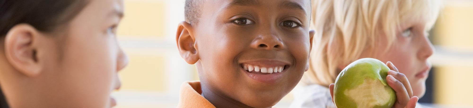 Kid smiles as he eats apple, othe kid stares at him.